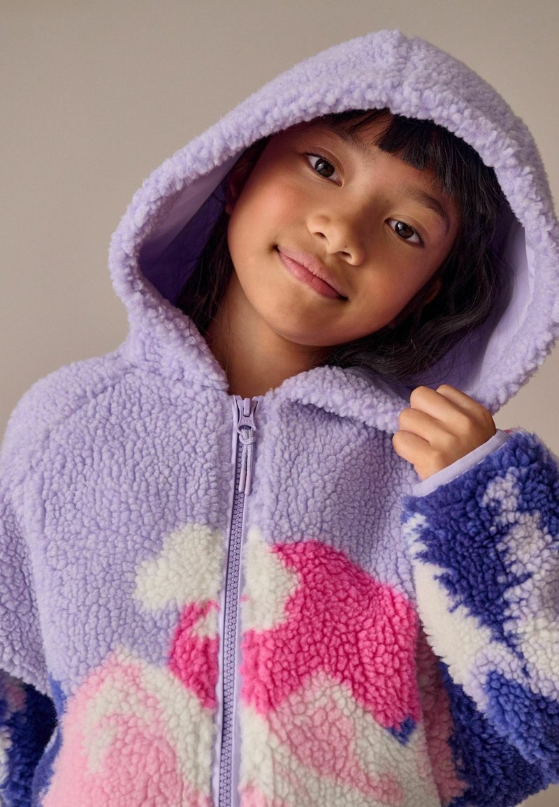 Haut en polaire violet avec fermeture éclair à l'avant, présentant des motifs abstraits en rose, blanc et bleu. Tissu texturé et grande capuche pour plus de chaleur.