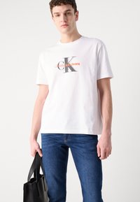 Hvit bomulls T-skjorte med et grafisk logo med "Calvin Klein Jeans" i svart og oransje. Kombinert med blå denim jeans og en svart veske.