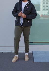 Czarna lekka kurtka z kapturem i białym zamkiem błyskawicznym, szara koszulka, oliwkowozielone spodnie oraz beżowe buty. Widoczna faktura na powierzchni obuwia.