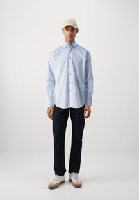 Hellblaues Hemd mit Kentkragen, getragen zu dunklen Jeans und einer beigen Mütze. Weiße Sneakers mit sandfarbenen Akzenten. Modell steht vor einem schlichten Hintergrund.