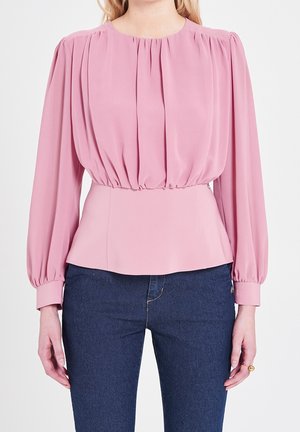 Nainen, joka pukeutunut pitkähihainen, laskostettu vaaleanpunainen pusero, jossa on istuva vyötärö, ja tummansiniset farkut, seisoo yksinkertaisen taustan edessä.