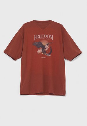 Homme portant un t-shirt marron rouille avec un motif d'aigle, un pantalon cargo coloré imprimé représentants des cavaliers et un paysage, des chaussures noires et un sac bandoulière.