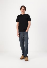 Ung man som står med en hand i fickan, iklädd en svart t-shirt, blå jeans och bruna vardagsskor mot en vit bakgrund.