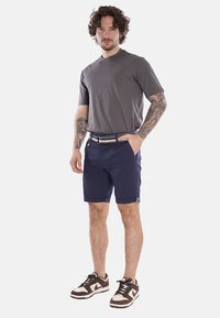 Maglietta a maniche corte grigio scuro abbinata a pantaloni corti blu, con una cintura a righe; il modello indossa scarpe da ginnastica marroni. Tessuto liscio, vestibilità casual.