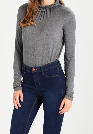 Woman wearing a fitted grey long-sleeve top with a gathered neckline and high-waisted dark blue jeans.