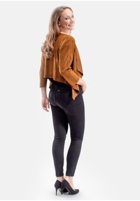 Bruine suède jas met een cropped ontwerp en wijde mouwen, gecombineerd met strakke zwarte jeans en zwarte hoge hakken.