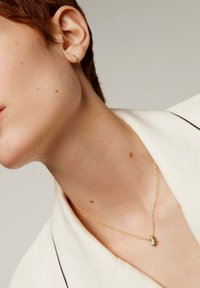 Collar de oro con un pequeño colgante de turquesa, combinado con un simple cuff en la oreja. Lleva puesto un prenda blanca con cuello. Textura de piel suave visible.