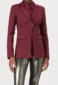 Blazer burdeos de diseño ajustado, solapas, dos bolsillos delanteros y un adorno redondo plateado. Combinado con pantalones metálicos de corte slim.