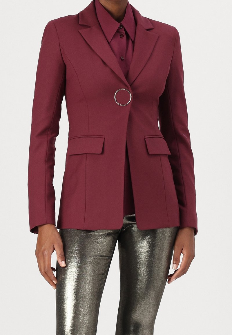 Blazer burdeos de diseño ajustado, solapas, dos bolsillos delanteros y un adorno redondo plateado. Combinado con pantalones metálicos de corte slim.