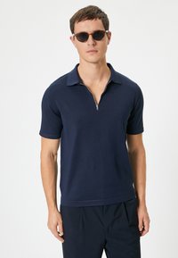 Marinblå kortärmad polo med dragkedja i kragen, ribbad textur, subtilt figurnära design, kombinerad med mörka solglasögon och byxor.