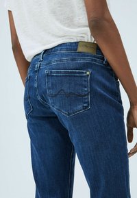 Osoba nosící tmavě modré džíny s prošíváním na zadních kapsách a malou koženou nášivkou na pásku, v kombinaci s bílou košilí.