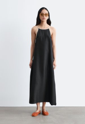 Femme portant une robe maxi noire sans manches avec un bord blanc, des lunettes de soleil ambre et des ballerines orange, debout contre un mur blanc uni.