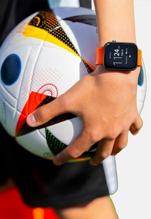 Un pallone da calcio bianco con motivi colorati è tenuto da una mano che indossa uno smartwatch nero con cinturino arancione, che mostra l'orario.