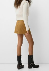 Beige plysch ärmlös väst, vit ribbad långärmad topp, beige mocka minikjol och svarta läderstövlar vid vadbenen med spännen.