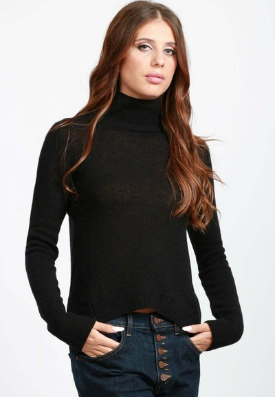 Giovane donna con lunghi capelli castani che indossa un maglione a collo alto nero e jeans blu, mani in tasca, espressione neutra.