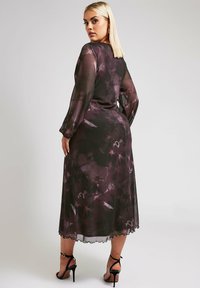 Przezroczysta, długa sukienka z rękawami, w kwiatowy wzór w głębokim purpurze i czerni, wykończona falbaniastym dołem. Noszona na czarnych sandałach na obcasie.