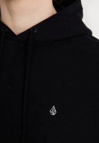 Zwarte sweatshirt met capuchon, voorzien van een klein wit geborduurd logo op de linkerborst. Gemaakt van zachte katoen met textuur aan de koordjes.