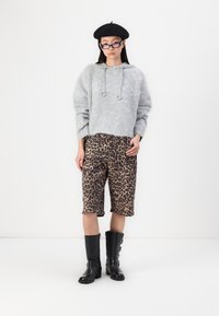 Ljusgrå, fluffig hoodie med dragsnören, i kombination med shorts med leopardmönster och knähöga svarta stövlar. Modellen bär en svart basker och glasögon.