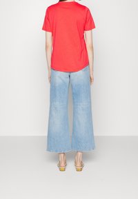 Person wearing a red short-sleeve shirt, light blue wide-leg jeans, and white heeled sandals facing away against a plain background.