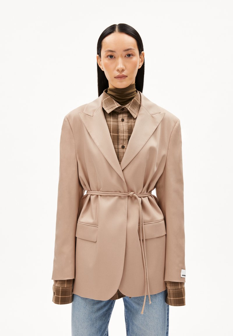 Blazer beige con vita legata, dotato di ampi risvolti e due tasche frontali, indossato sopra una camicia a quadri marrone. Jeans blu completano l'outfit.