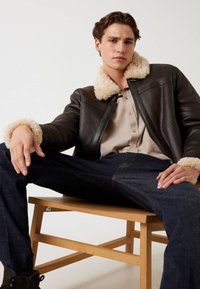 Giacca di pelle marrone con colletto in shearling beige, camicia chiara a bottoni e jeans in denim scuro, seduto su una sedia di legno.