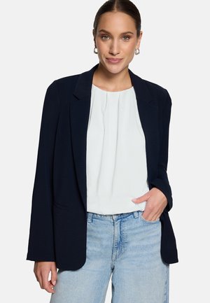 Frau mit silbernen Creolen, weißer Bluse, schwarzem Blazer und hellblauen Jeans, posierend mit einer Hand in der Tasche vor weißem Hintergrund.