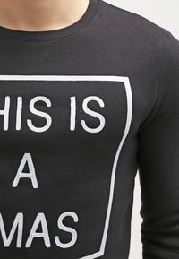 Czarna bluza z białym haftowanym napisem w prostokątnej ramce "TO JEST A". Zbliżenie na teksturę materiału i dekolt.