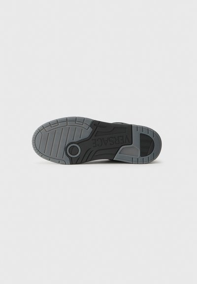 Black sneaker sole with textured rubber, featuring tread patterns and a circular accent. "VERSACE" logo embossed on the side.
