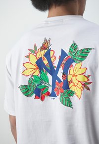 Camiseta de manga corta blanca con un diseño floral colorido y un gran logo azul de “NY”, resaltado por flores rojas y amarillas y hojas verdes.