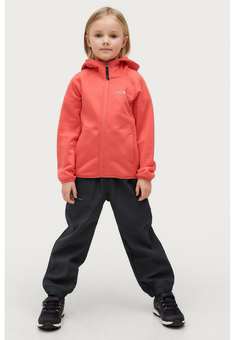 Koraalroze ritsvest met een zachte textuur, gecombineerd met losse zwarte broek en zwarte sportschoenen. Twee voorzakken zichtbaar.