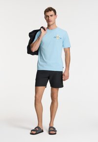 Himmelblaues Baumwoll-T-Shirt mit Grafikdruck, kombiniert mit schwarzen Shorts und dunklen Sandalen. Das Modell hält eine schwarze Jacke über die Schulter.