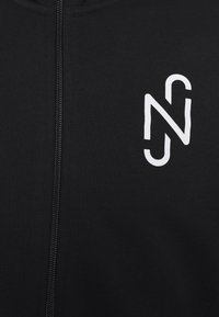 Hoodie à fermeture éclair noir en tissu doux, présentant un logo stylisé blanc sur la poitrine à gauche. Texture lisse, coupe ajustée, sans détails supplémentaires.