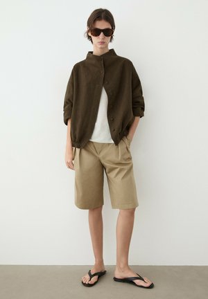 Personne portant une veste boutonnée marron foncé oversize, un haut blanc, un short beige longueur genou, des sandales noires et de grandes lunettes de soleil foncées, se tenant contre un mur blanc.