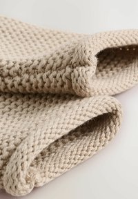 Tissu en coton tricoté beige avec un motif texturé et tissé, présentant des bords roulés. Le matériau semble doux et épais.
