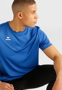 Blauwe sportieve T-shirt gemaakt van glad, vochtafvoerende stof. Heeft een ronde hals en een klein wit logo op de linkerkant van de borst.