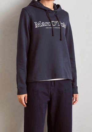 Osoba nosící tmavě modrou mikinu Marc O'Polo s tkaničkami a volné tmavé kalhoty stojí opřená o jednoduchou světlou zeď.