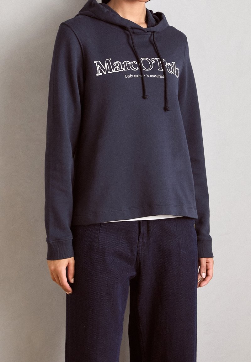 Personne portant un sweat à capuche bleu marine Marc O'Polo avec cordons et un pantalon large foncé, debout contre un mur clair et uni.