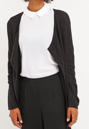 Personne portant une chemise blanche à col, un cardigan noir texturé ouvert sur le devant, et un pantalon noir taille haute sur un fond uni.