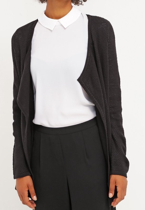 Personne portant une chemise blanche à col, un cardigan noir texturé ouvert sur le devant, et un pantalon noir taille haute sur un fond uni.