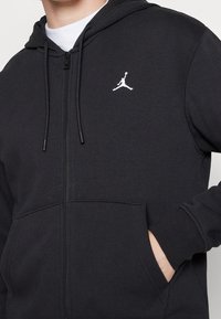 Uomo che indossa una felpa nera con zip e logo Jumpman bianco sul petto, con una mano nella tasca anteriore, sopra una maglietta bianca.