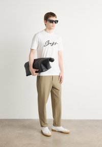 Fehér póló "Boss" felirattal, bézs nadrág, fekete kézitáska, napszemüveg és fehér sneakers barna talppal. Minimalista stílus.
