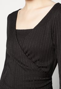 Top noir à manches longues avec un décolleté carré, texture côtelée et design croisé, mettant en valeur une silhouette ajustée.