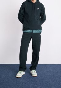 Mörkgrön fleecehoodie med en ficka fram och justerbara dragsnöre, i kombination med matchande fleecebyxor och ljusgröna sneakers.