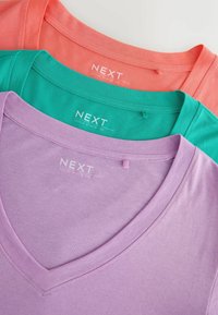 Trois T-shirts à col en V soigneusement empilés dans des tons pastel corail, sarcelle et lavande, chacun portant l'étiquette "NEXT Slouch V-neck Tee" à l'intérieur du col.