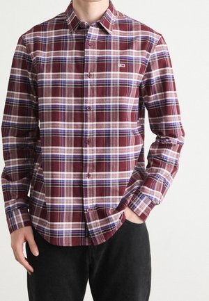 Homme portant une chemise à carreaux bordeaux, blanche et bleue, à manches longues avec col, et un pantalon noir.
