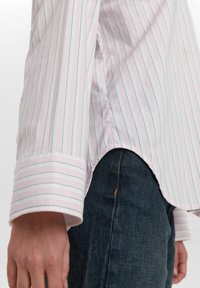 Light pink striped shirt with short sleeves, featuring light blue and white stripes, curved hem, and a smooth fabric texture. Paired with dark denim.