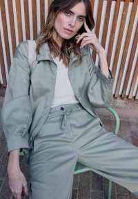 Femme aux cheveux châtain clair et aux yeux verts, portant une veste et un pantalon vert clair, assise sur une chaise verte devant un mur en panneaux de bois.