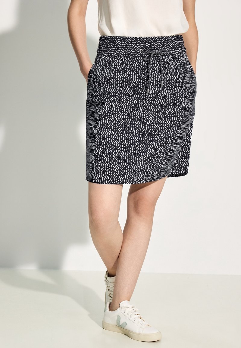Jupe marine avec motif de vagues blanches, taille élastique avec cordon de serrage et poches latérales. Associée à des baskets blanches avec des accents verts.