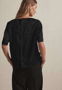 Black short-sleeve top with a textured surface, straight neckline, and back seam detail. Paired with black pants and simple accessories.