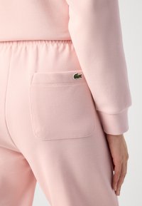 Ροζ κωδικοί φόρμες με υφή διαθέτουν ελαστική μέση και μια πίσω τσέπη με το πράσινο λογότυπο της Lacoste. Κοντινή λήψη της πίσω λεπτομέρειας.